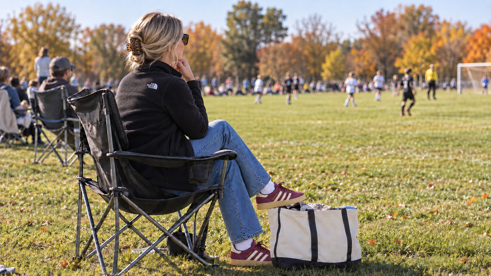 Blonde mother with her hair in a claw clip, black fleece and burgundy sneakers watching a fall soccer game from a sideline chair